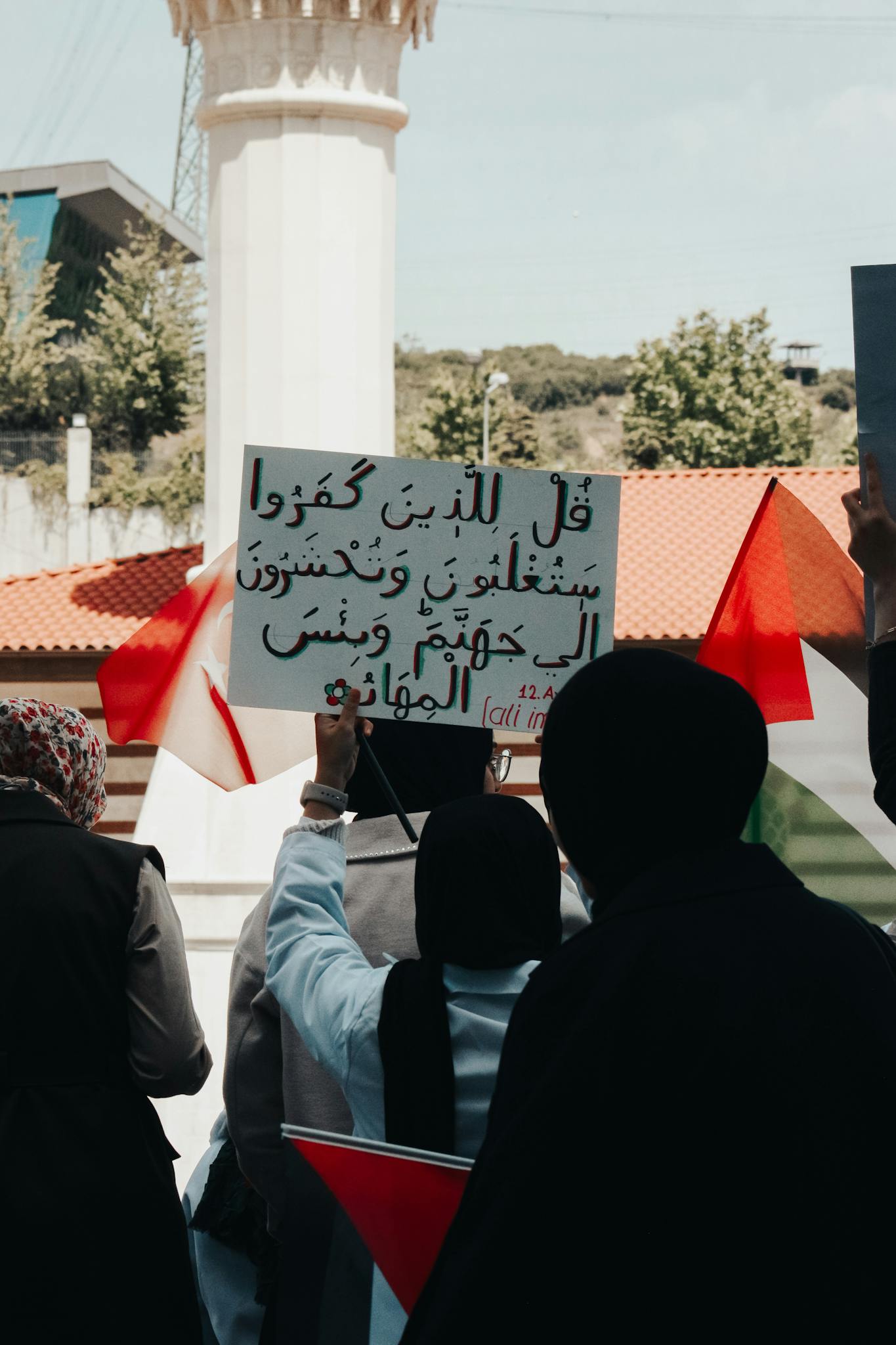 Women in hijabs hold a sign and Palestinian flags in a peaceful street demonstration.