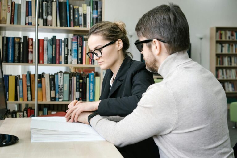 A caregiver helps a visually impaired man read a Braille book in a library setting.
