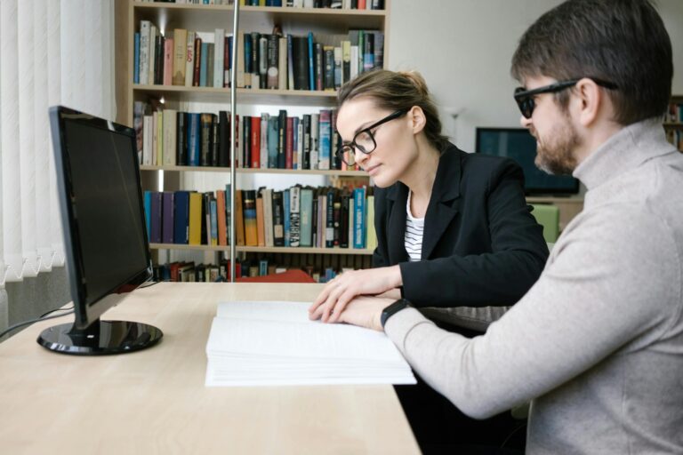 A caregiver helps a visually impaired man read a braille book in a library setting.